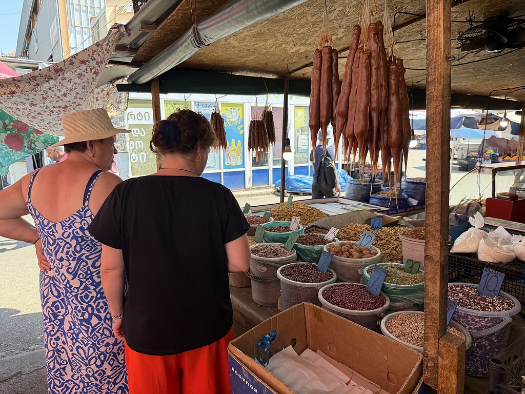 Zwei Damen am Marktstand auf dem Dezerter Bazaar in Tiflis mit hängenden Tschurtschchela, Eimern voller Nüsse und Bohnen und handgeschriebenen Preisschildern