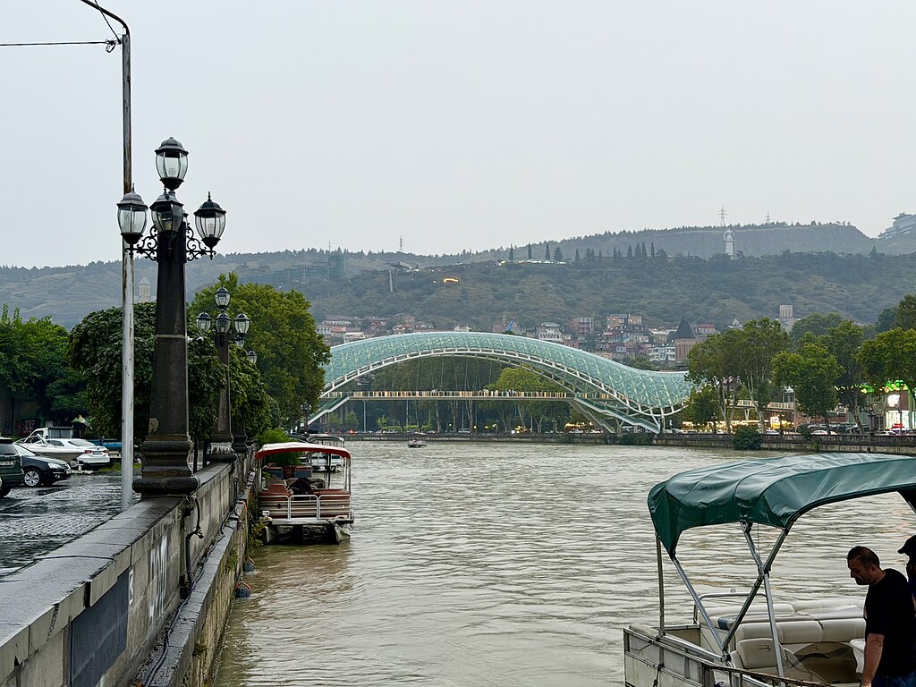 Friedensbrücke Tiflis über den Fluss Kura. Es fahren Boote auf dem Fluss und auf der Ufer Mauer stehen Laternen