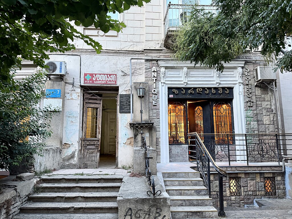 Zwei Hauseingänge nebeneinander in Tiflis, Georgien. Links und rechts stehen Bäume. Zu den Türen führen Treppen. Beide stehen offen. Dabei sieht die linke Haushälfte wesentlich älter und im Baufälligerem Zustand aus als die rechte Haushälfte. Diese ist neuer