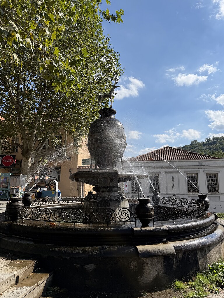 Brunnen in Sighnaghi auf einem PLatz. Er ist mit den georgischen Weintonfässern Qvevri verziert. In der Mitte steht ein Hirsch auf einem Qvevri. Im Hintergrund stehen Häuser und es ist blauer Himmel.