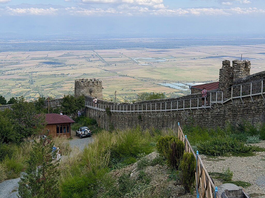 Stadtmauer mit zwei Türmen in Sighnaghi. Sie verläuft am Hang umgeben von grünen Wiesen, Feldern und einem HAus. Neben dem Haus parkt ein Auto. Der Himmel ist blau mit Wolken.