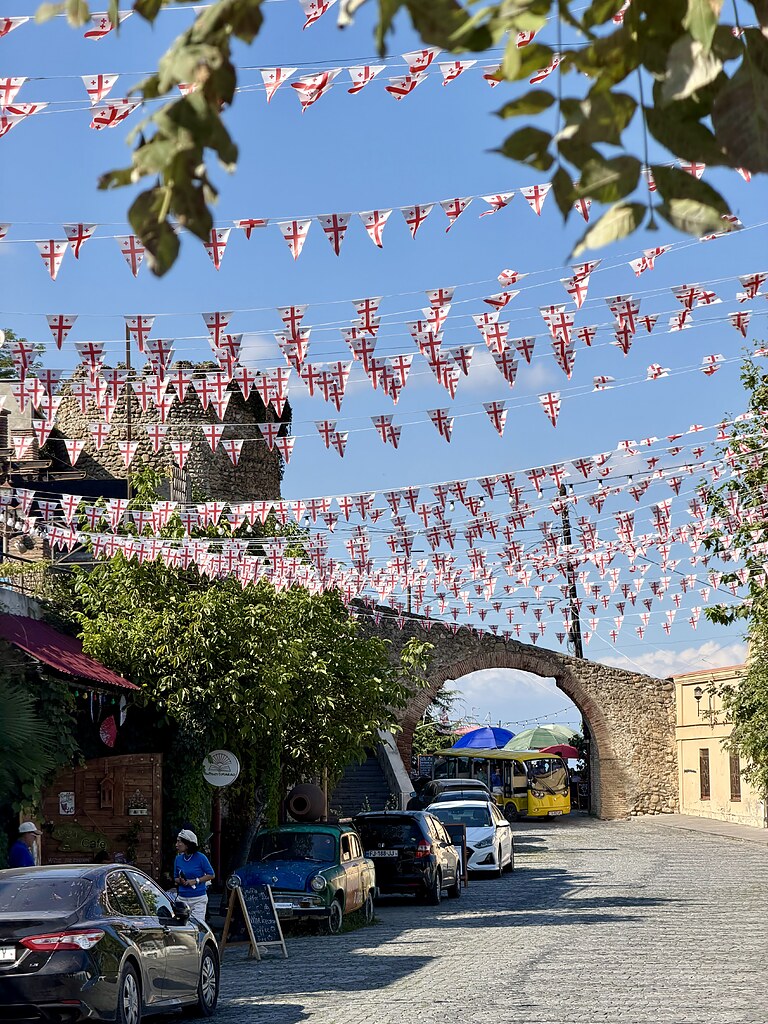 Wimpel mit der georgischen Flagge hängen über eine Straße in Sighnaghi, es führt ein Bogen der Stadtmauer über die Straße. Darunter fährt ein gelber Touristenbus entlang. An der linken Straßenseite parken Autos. Die Sonne scheint und der Himmel ist blau