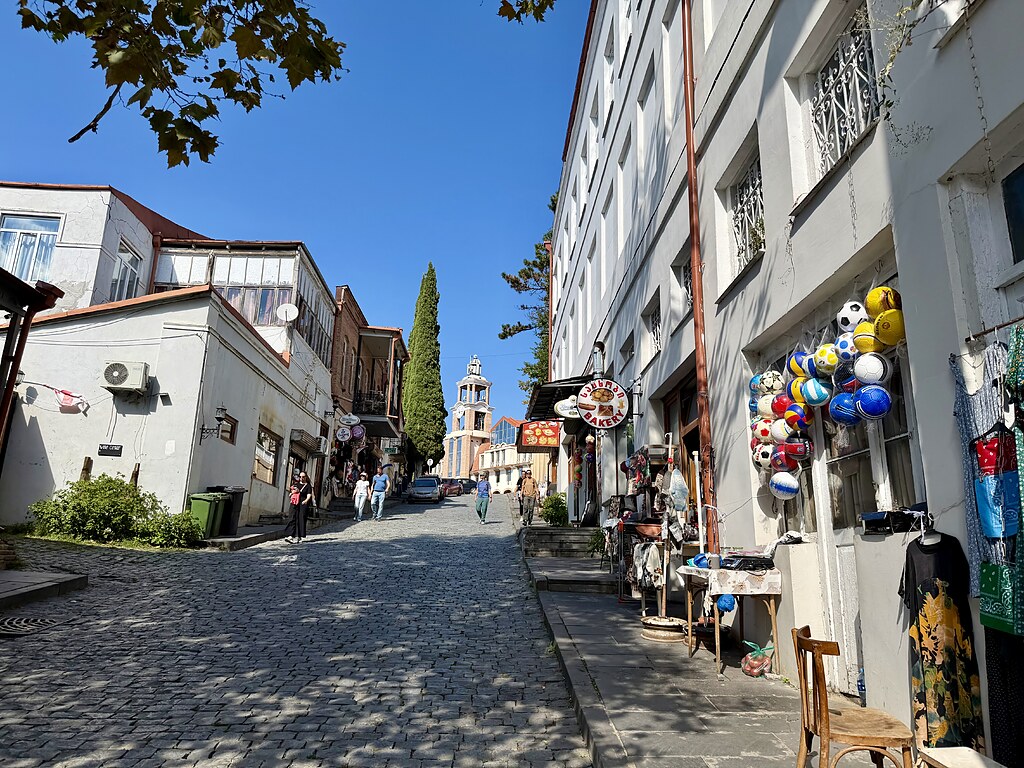 Straße in Sighnaghi, mit einem Bäcker und verschiedenen Läden an der Straße. Es geht den Berg hoch, oben auf dem Berg steht ein Turm mit einer Uhr.