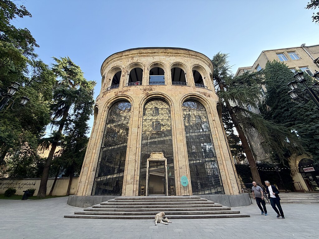 Ein imposantes Gebäude in Tiflis., Georgien. Hier Starten die Gondeln zum Mtatsmindapark. Vor dem EIngang ist ein grünes Schild mit der Aufschrift „Tickets“. Vor dem Eingang befinden sich Treppen, wovor ein brauner Straßenhund liegt. Rechts und links neben dem Gebäude stehen Tannenbäume