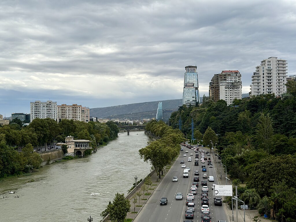 Links der Fluss Kura, rechst eine mehrspurige Straße mit vielen Autos. Weiter hinten führt eine Brücke über den Fluss und es stehen Hochhäuser vor einer Bergkette. Rechts sind noch viele Bäume. In Tiflis, Georgien