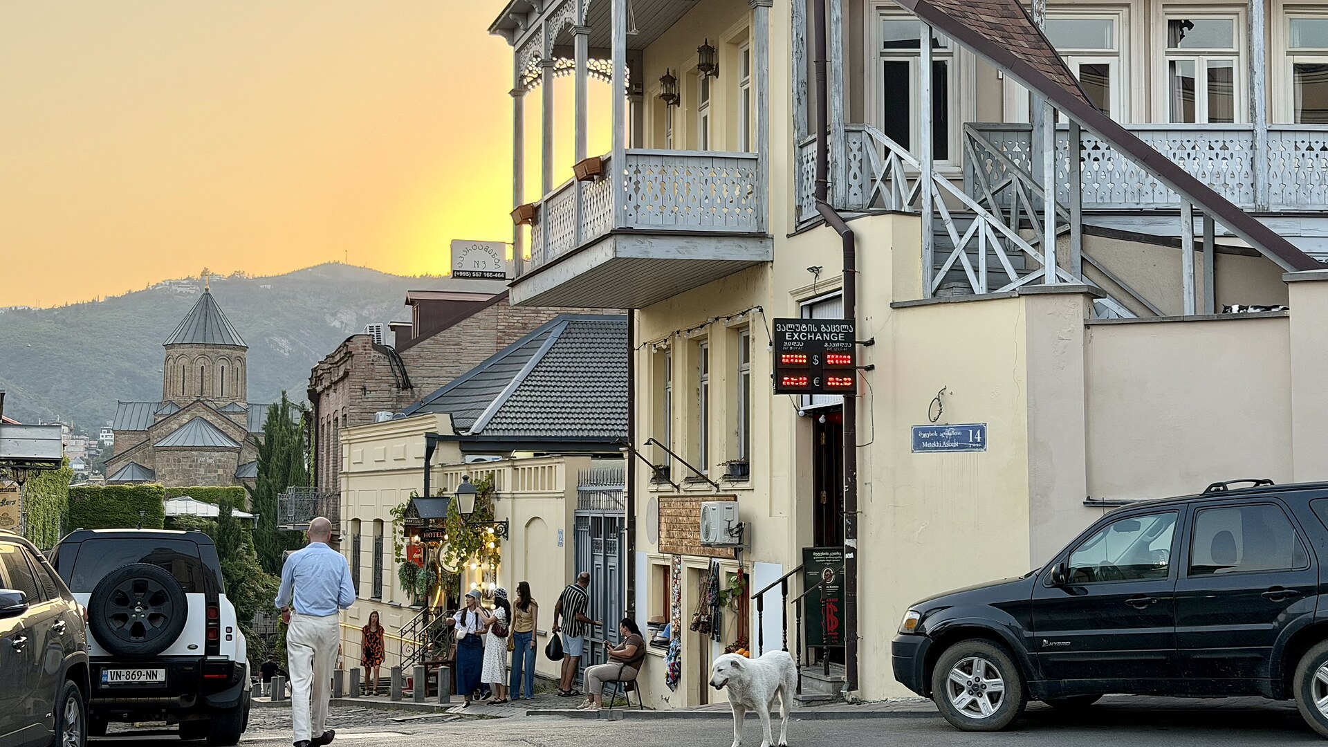 Gelbe Hausfassaden mit hellblauen, verzierten Balkonen, einem Schild einer Wechselstube und verschiedenen Eingänge, in einer Straße die Bergab in Tiflis, Georgien. Auf der Straße steht ein Hund, es laufen und sitzen ein paar Menschen und es parken Autos an der Seite. Weiter hinten steht eine Kirche mit einem spitzen Dach und im Hintergrund sind Berge und ein Sonnenuntergang.