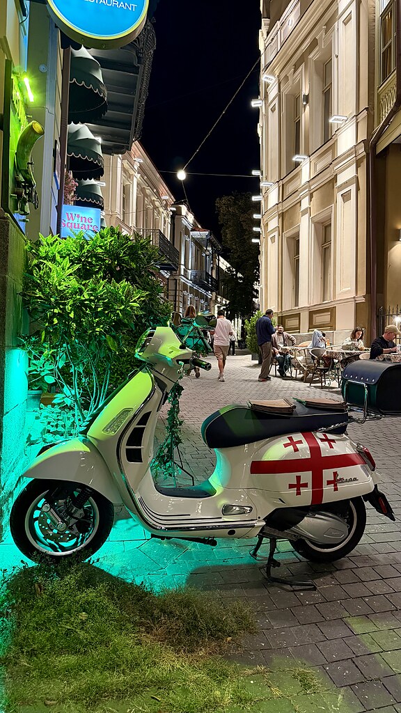 Weißer Motorroller mit Speisekarten auf dem Sitz und der Georgischen Flagge aufgedruckt steht in einer beleuchteten Altstadt Gasse in Tiflis in der Menschen an Tischen und Stühlen sitzen und essen.