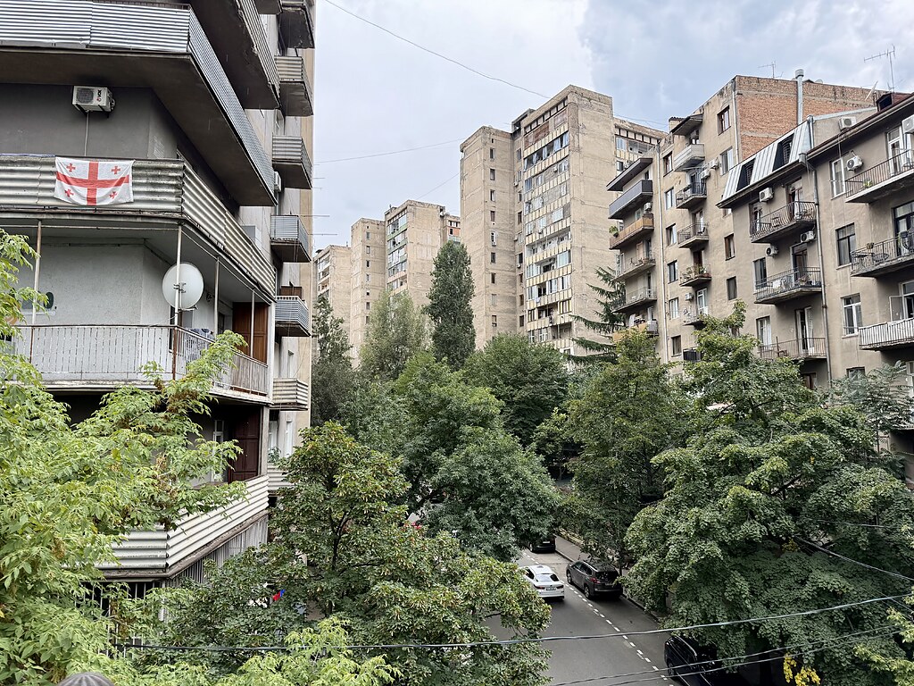 Alte, Ostblöcke aus der Sowjet Zeit in Tiflis, Georgien, dazwischen eine Straße wo Autos parken und Bäume. An einem Balkon hängt eine georgische Flagge.