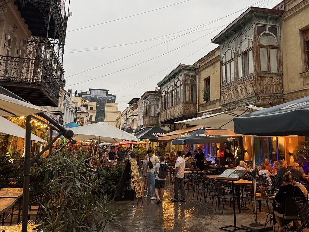 Eine Straße mit vielen Restaurants, aufgeklappten Sonnenschirm und Holzbalkonen an den Fassaden. Es sind viele Menschen dort in der Altstadt von Tiflis, Georgien