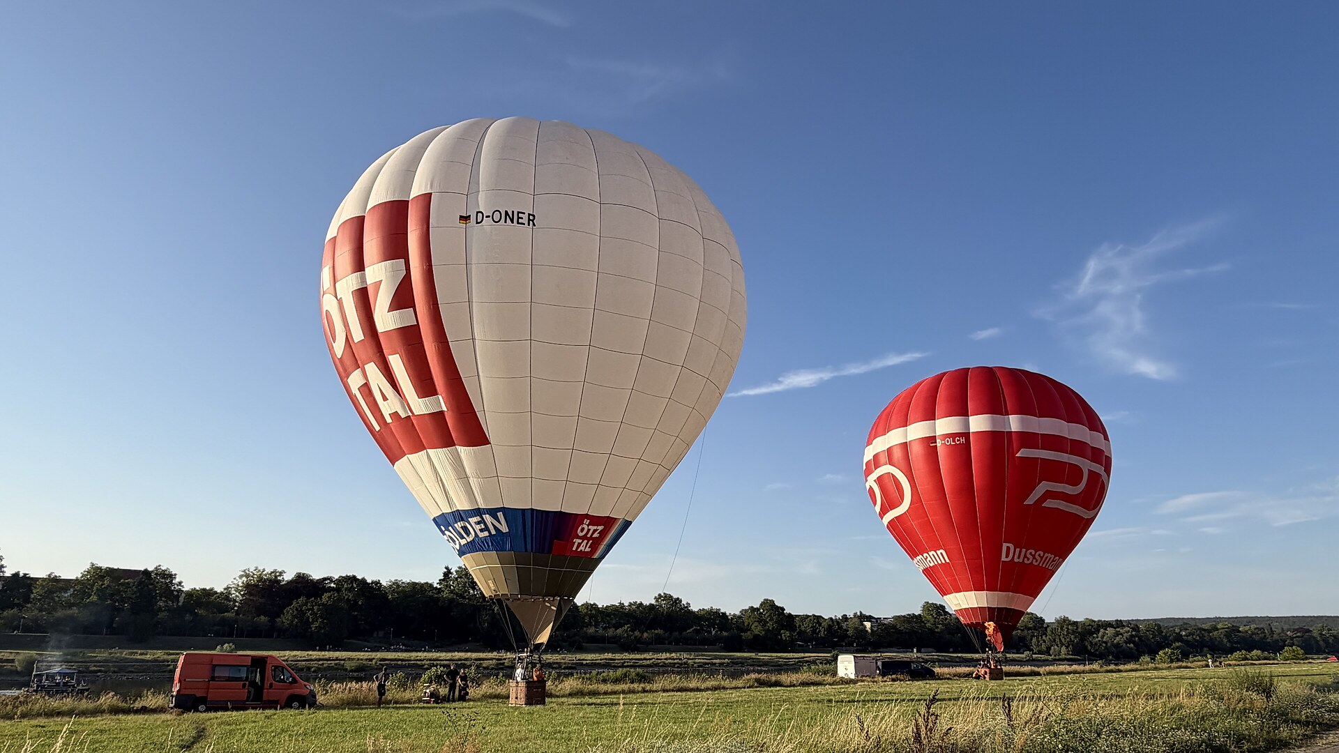 Ein roter und ein weißer Ballon, fertig zum Start auf einer grünen Wiese am Ufer von der Elbe, in Dresden. Daneben ist ein Orangener Bus und ein schwarzer Bus mit weißem Anhänger.