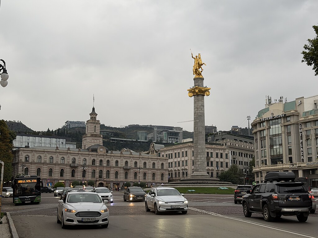 Die Statue vom Liberty Square steht in der Mitte vom Kreisverkehr. Drum herum fahren viele Autos und ein Bus. Tiflis, Georgien