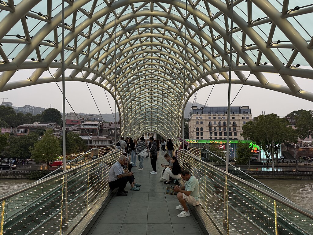 Auf der beleuchteten Friedensbrücke in Tiflis warten Menschen das es aufhört zu regnen. Sie sitzen an den Seiten der Brücke