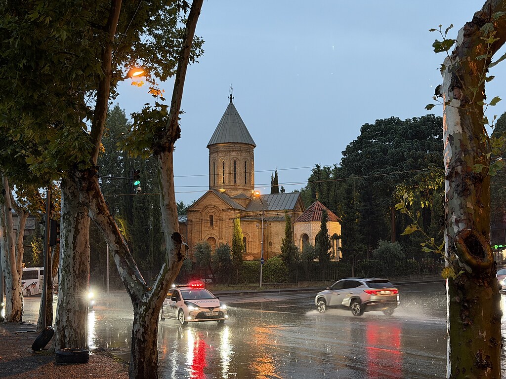 Eine kleine Kirche an der Straße neben dem Fluss Kura in Tiflis, Georgien. Davor fährt ein Polizeiauto bei Regen.