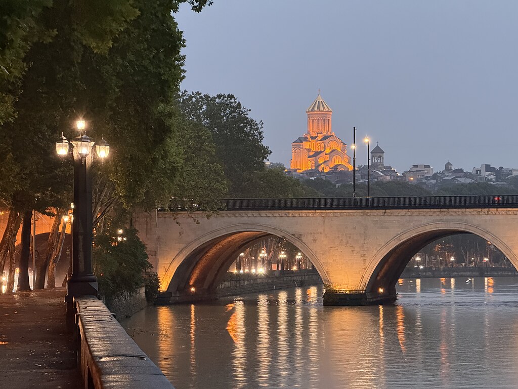 Blick vom Fluss Kura auf die Sameba-Kathedrale, leuchtende Straßenlaternen und eine Brücke in Tiflis bei Regen