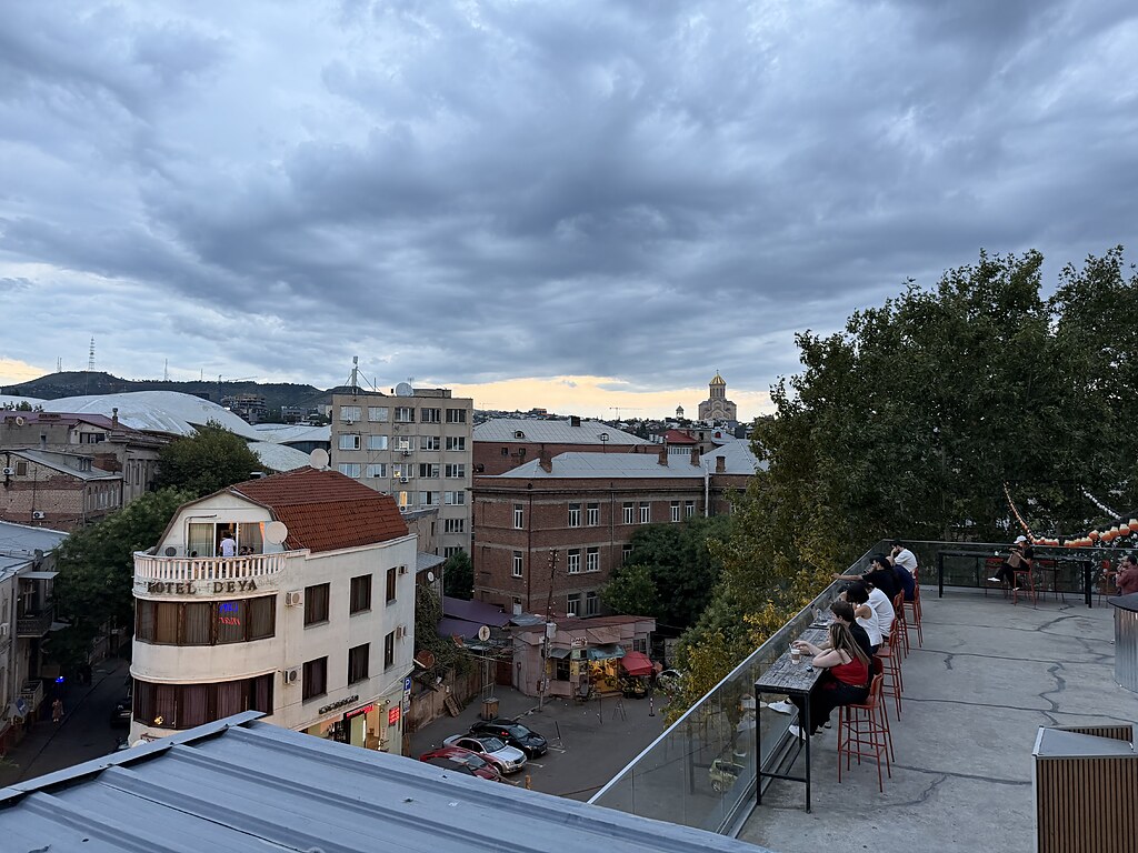 Dachterasse vom Orbeliani Bazaar in Tiflis, Georgien. Oben sitzen Menschen mit Getränke und schauen auf die Altstadt FAssaden. In weiter Ferne sieht man die Sameba-Kathedrale.