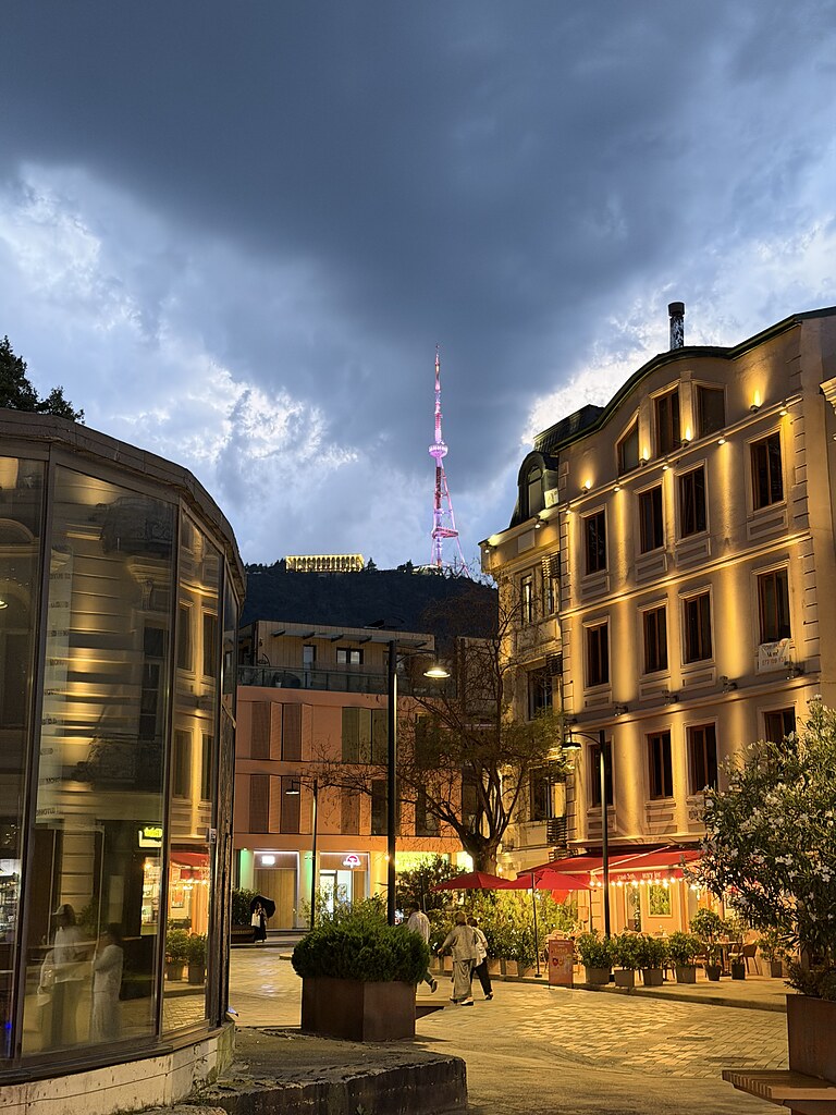 Beleuchtete Altstadt Häuser von Tiflis, Georgien. Davor rote Schirme mit Lichterkette und Menschen. Oben auf dem Berg, steht der Fernsehturm.