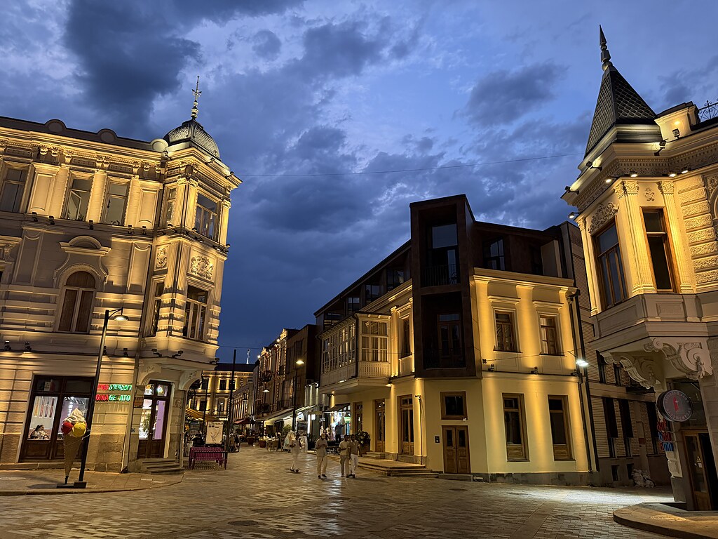 Verzierte Häuserfassaden mit einem spitzen Turm, und Gassen in der Altstadt von Tiflis, Georgien. Die Häuser Erden im dunkeln beleuchtet. Ein paar Menschen sind auf den Straßen.