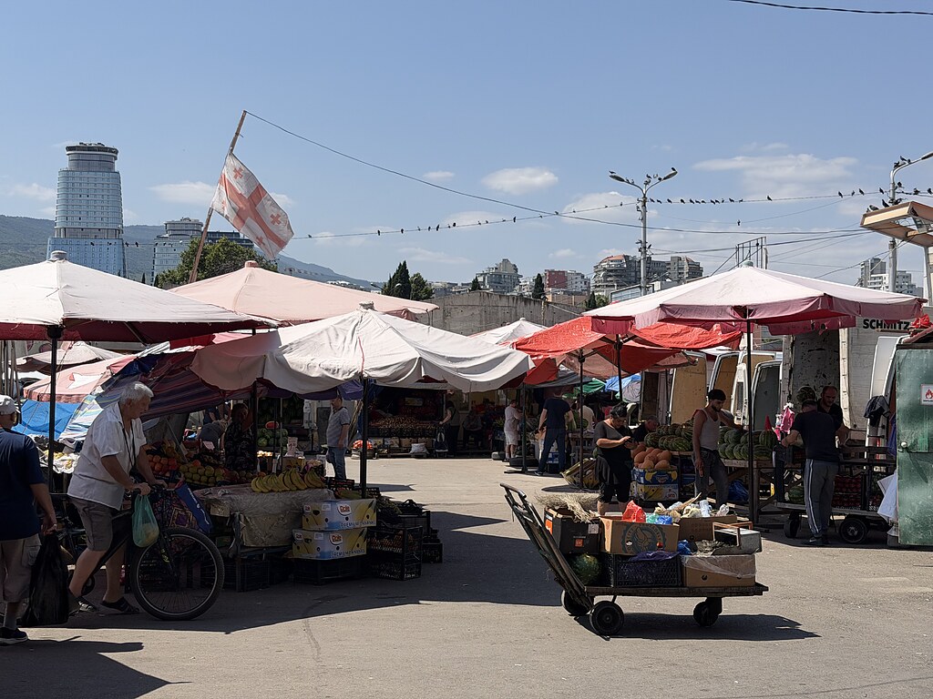 Gemüsestände, Obst Karren und eine Georgische Fahne auf dem Dezerter Bazaar Tiflis. Dahinter sieht man Hochhäuser und viele rote Sonnenschirme