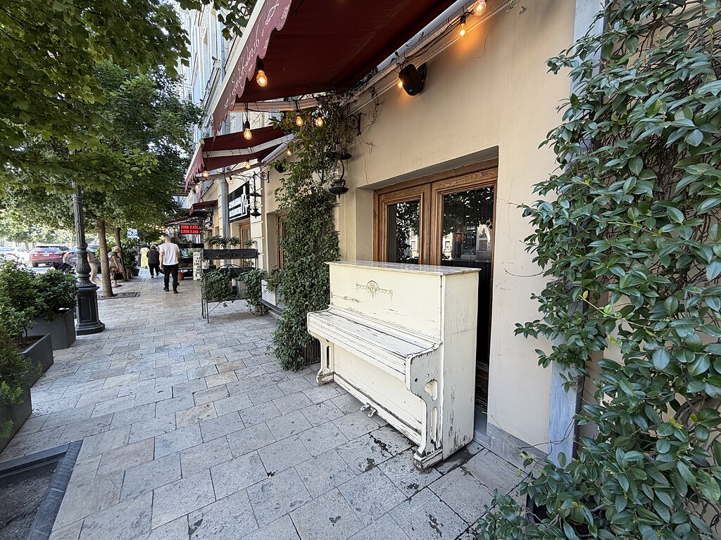 Ein Klavier vor einem Restaurantfenster auf einem Bürgersteig in Tiflis, Georgien. Da drüber ist eine rote Markise mit einer Lichterkette.