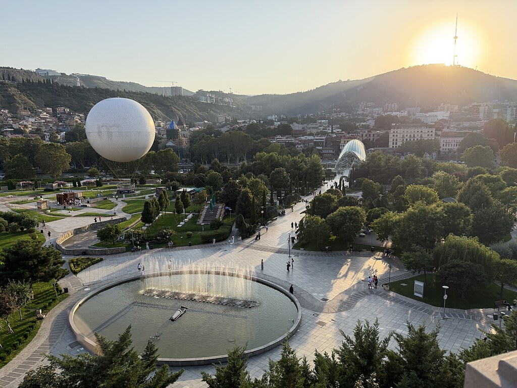 Rike Park in Tiflis, Georgien, ein Wasserbecken mit Fontänen, ein weißer Ballon, die Friedensbrücke im Grünen. Dahinter ein Bergpanorama, der Fernsehturm und Sonnenuntergang.