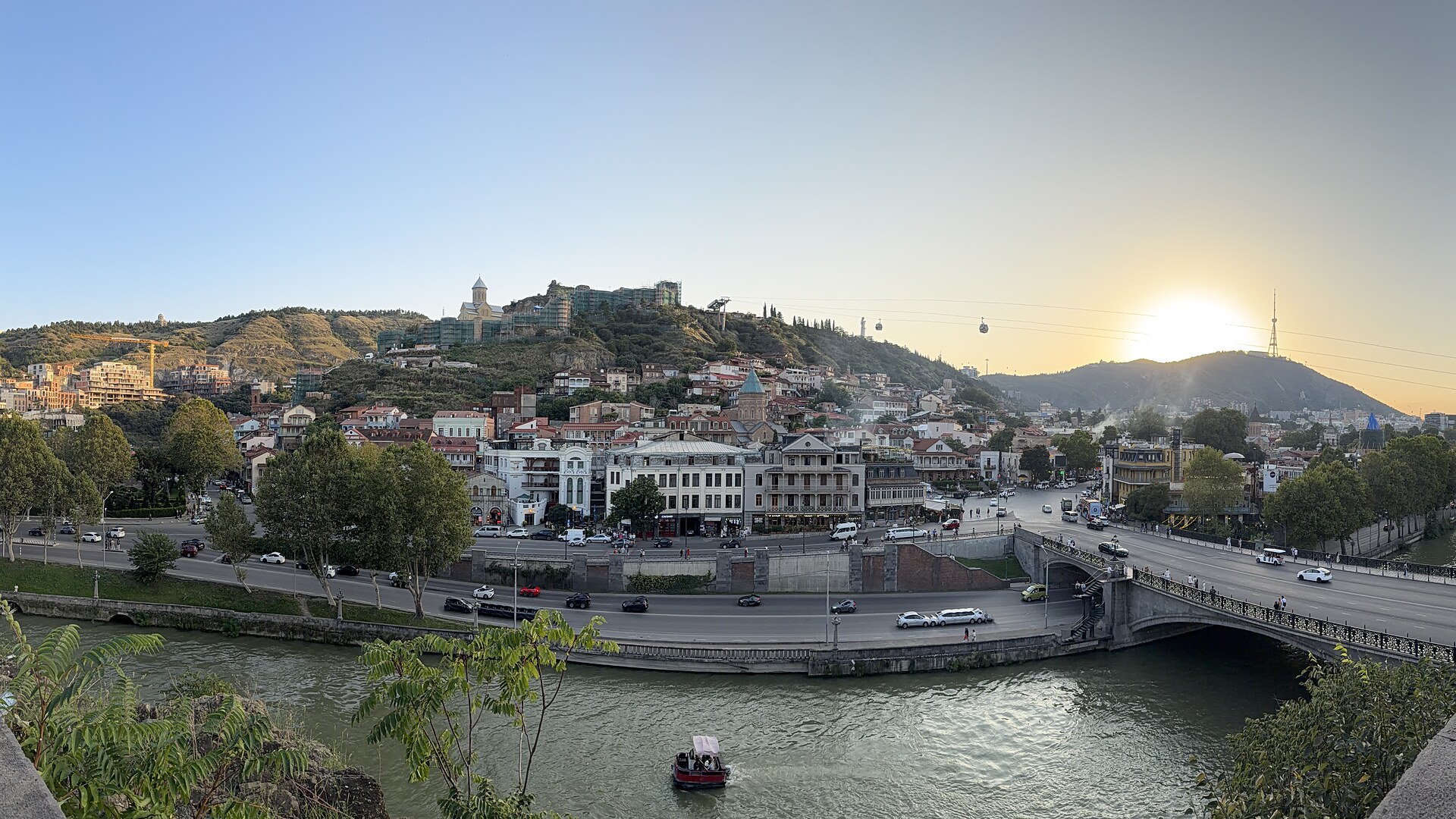 Panorama Ausblick über den Fluß, auf die Stadt Tiflis am Berg, bei einem Sonnenuntergang. Rechts im Bild ist eine Pferdestatue mit einem Reiter auf der Mauer