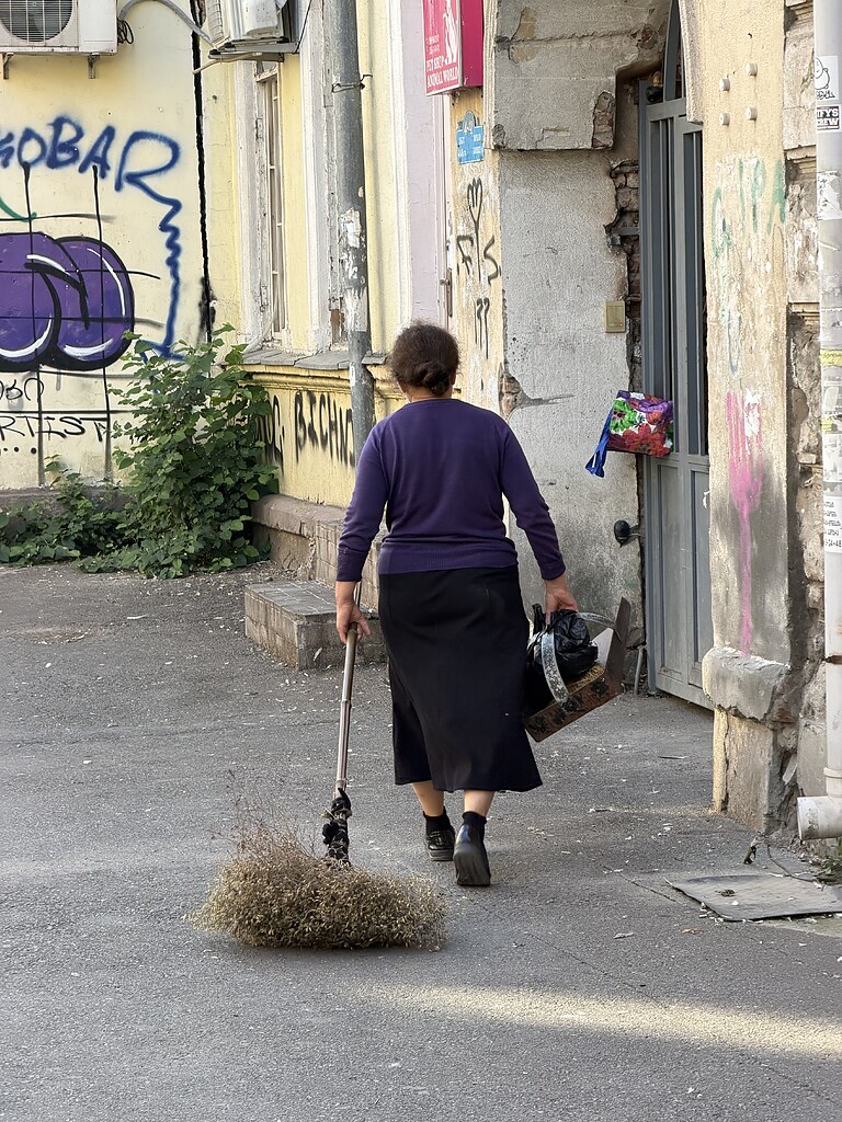 eine alte Dame mit einem Besen aus Naturmaterialien und einer Kehrschaufel, läuft zu einem Hauseingang in Tiflis, Georgien. Die Fassade von dem Haus ist mit Grafitti besprüht