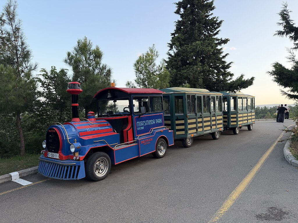Blau, rote Eisenbahn, Bimmelbahn mit zwei grün gelben Wagongs, dahinter stehen grüne Bäume, auch ein Tannenbaum