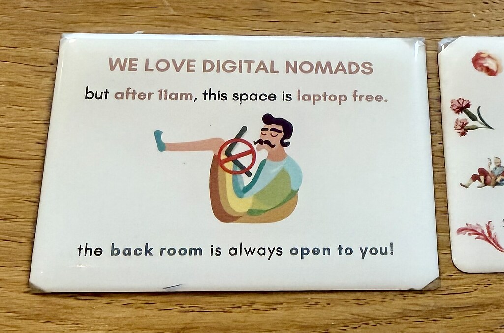 Schild auf einem Holztisch bei Shavi Coffee Roasters. Darauf ist ein Mann mit Schnurrbart mit einem Laptop über dem ein Verbotsschild gemalt ist. Es sagt, sie lieben digitale Nomaden, aber ab 11 Uhr ist der Bereich vorne Laptop frei