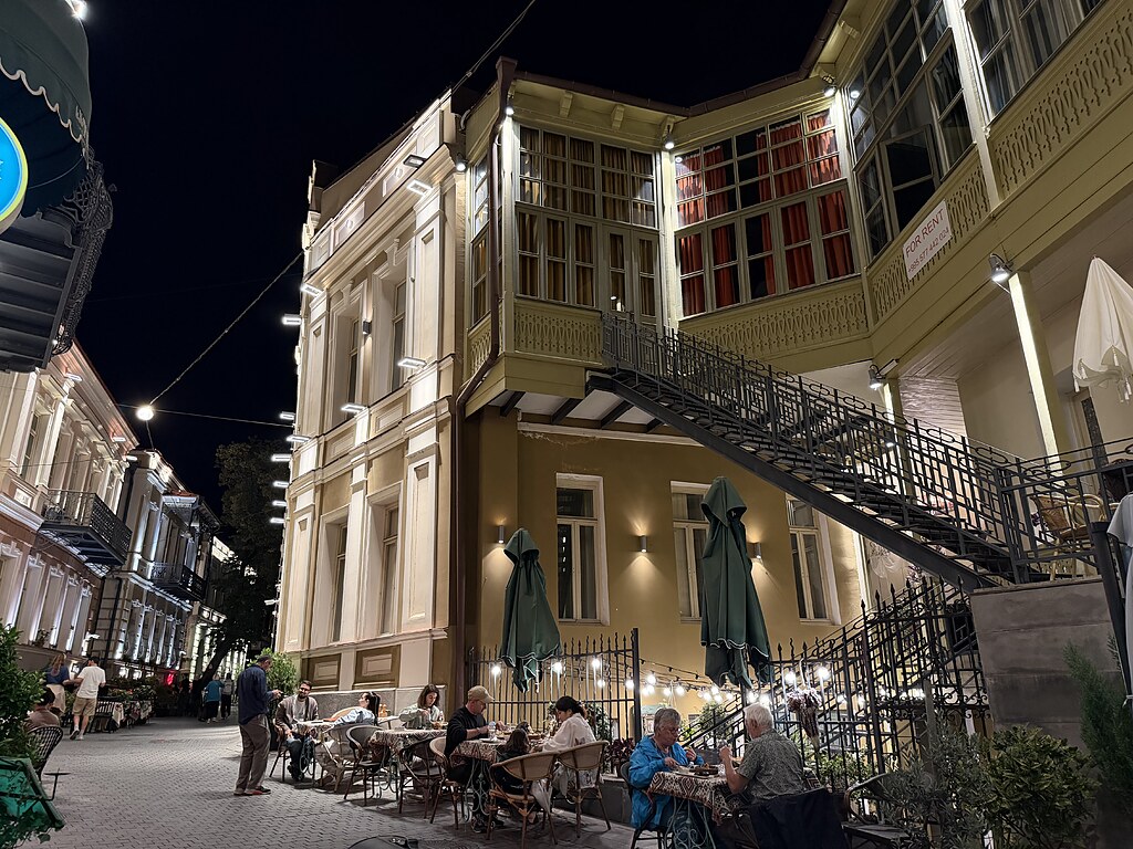 Altes, renoviertes Haus mit gelber Fassade und einer Metalltreppe außen. Davor sitzen Menschen an Tischen und Stühlen und essen. Es ist dunkel und die Gasse ist schön beleuchtet mit Lampen und Lichterketten. Tiflis, Georgien
