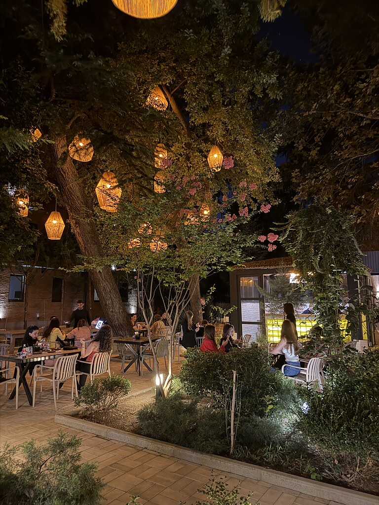 Bäume mit leuchtenden Lampions von einem Restaurant in einem Park in Tiflis, Georgien. Darunter sin Tische und Stühle und es sitzen Leute dort und essen.