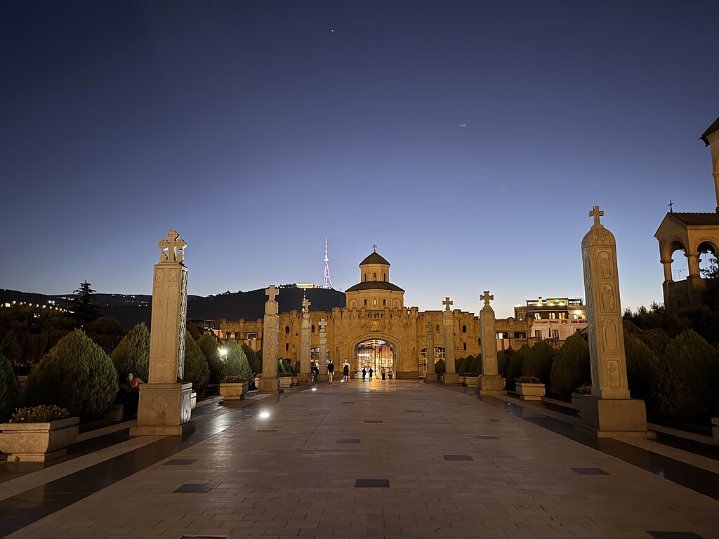 Prachtvoller Eingang zur Sameba-Kathedrale, beleuchtet in Tiflis, im Hintergrund steht der Fernsehturm beleuchtet im Mtatsminda Freizeitpark auf dem Berg