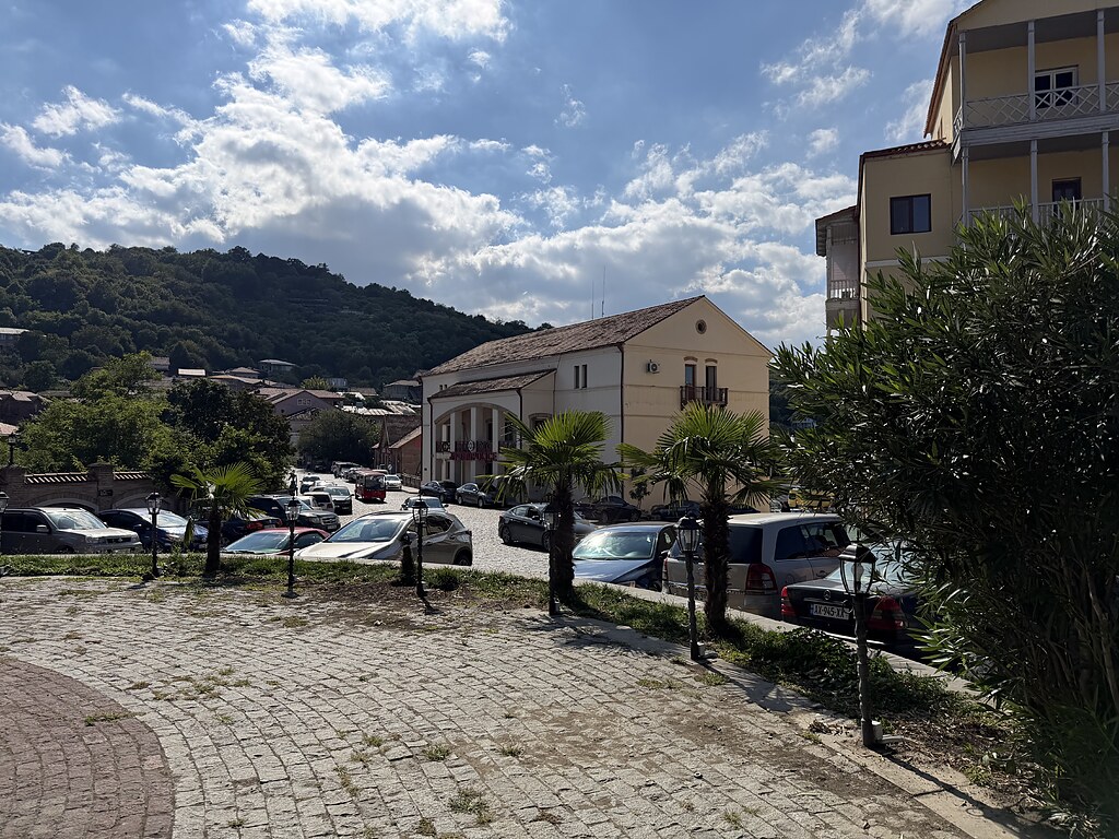 Ein Platz mit Palmen, dahinter parken Autos und es steht dort ein Polizeigebäude neben einem anderen Haus. Im Hintergrund stehen Berge und es gibt blauen Himmel mit Wolken.