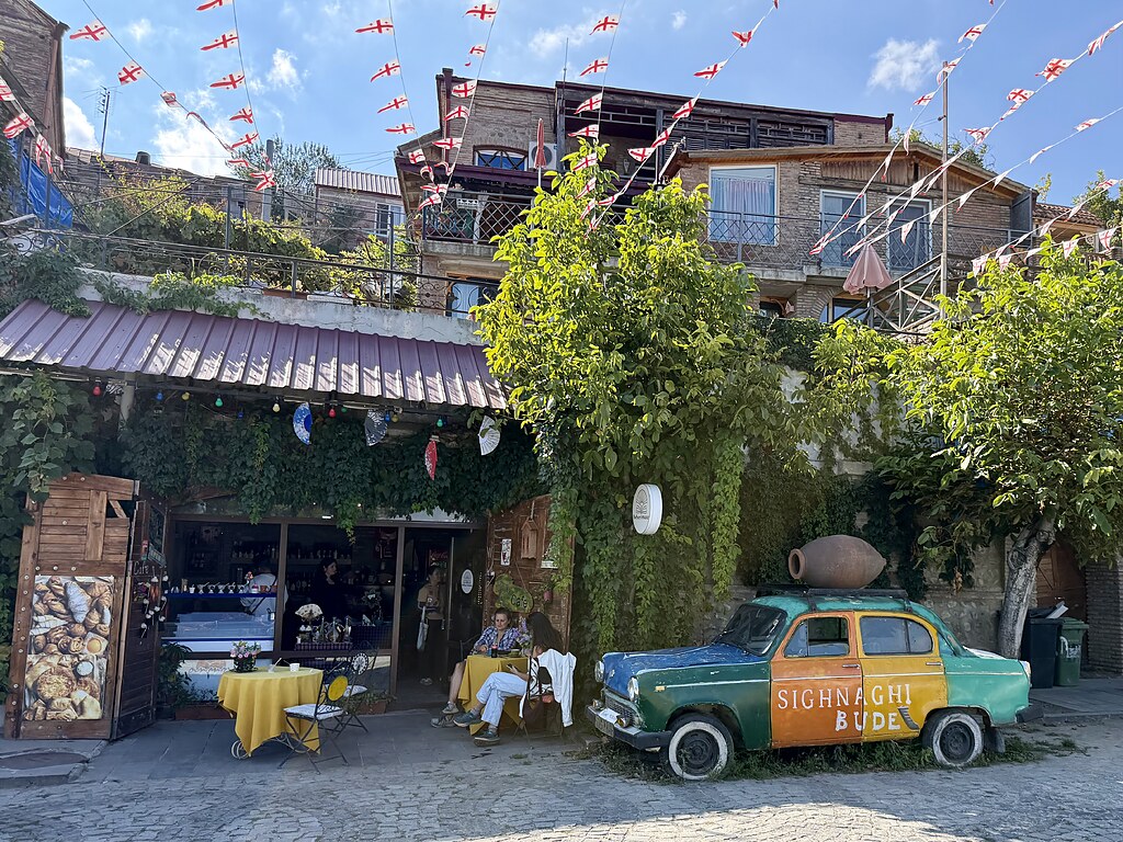 ein buntes Auto mit einem georgischen Ton Fass für Wein auf dem Dach neben einem Café in einer Häuserwand. Vor dem Cafe ist eine Straße über die Wimpel mit der georgischen Flagge gespannt sind.