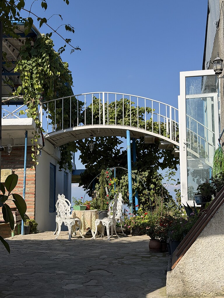 Ein weißer Metalöltisch mit zwei weißen Metallstühlen im gepflasterten Innenhof. Darum ranken Blumen und darüber eine kleine Metallbrücke in Form eines Bogens. Sie verbindet zwei Häuser miteinander. Der Himmel ist blau