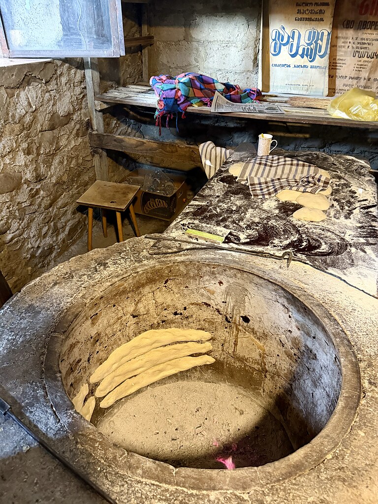 Shota Puri Ofen mit Brot, daneben frischer Brotteig auf einer Arbeitsfläche. DAneben ist ein altes Holzregal, eine Tasse und ein alter Holzhocker, in Georgien, Kachethien