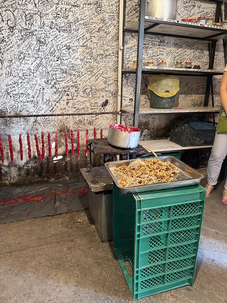 Eine Kiste mit einem Blech, auf dem aufgefädelte Walnüsse an Fäden liegen. Dahinter steht ein alter Topf mit einem Holzstab und einer roten Fruchtmasse. Dahinter an einer vollgeschriebenen Wand ist eine Stange auf der die frisch hergestellten Tschurtschchela hängen. Daneben steht ein Regal mit Gläsern, Eimern und einer Kiste an der Wand.