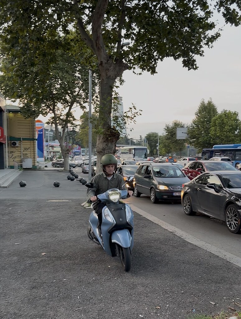 Essen Lieferant auf einem Moped fährt neben einer vollen Straße auf dem Bürgersteig in Tiflis, Georgien