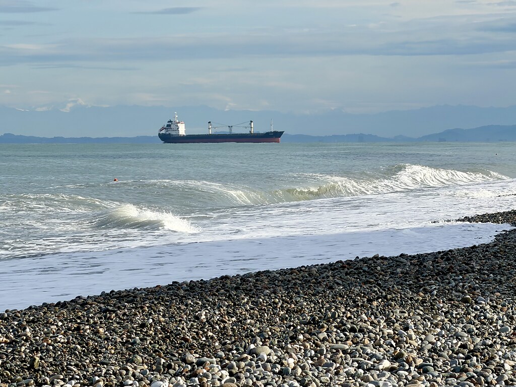 Schiff auf dem schwarzen Meer mit Wellen und Bergpanorama im Hintergrund, sowie ein Steinstrand in Batumi, Georgien.