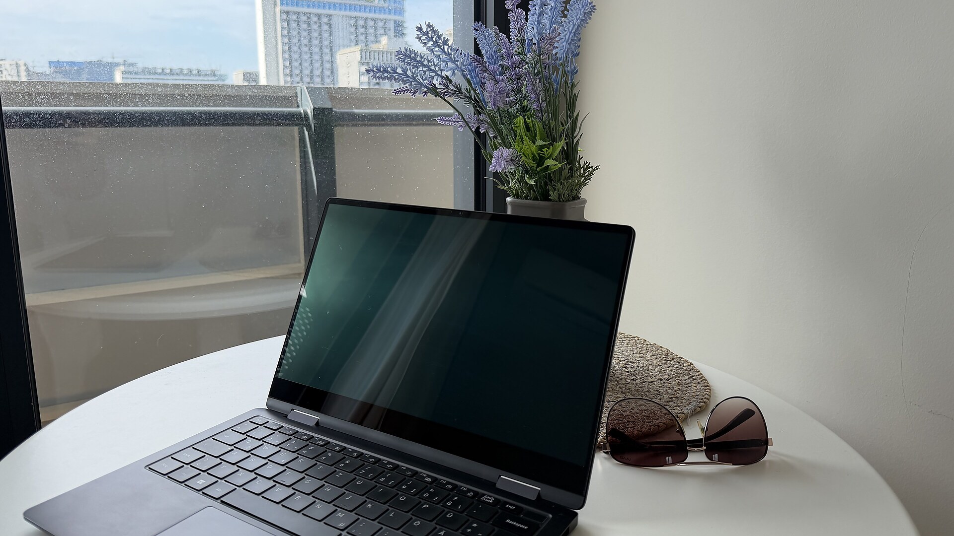 Laptop und Sonnenbrille liegen auf einem Tisch mit Blumen. Aus dem Fenster sieht man Hochhäuser in Batumi, Georgien.